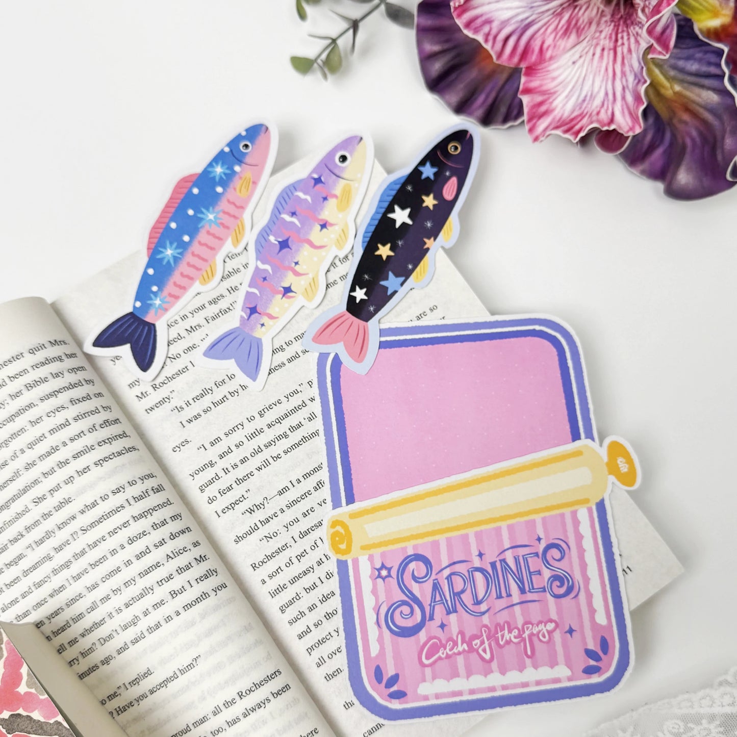 Three colorful sardine bookmarks removed from their pink Catch of the Page tin-shaped holder on an open book