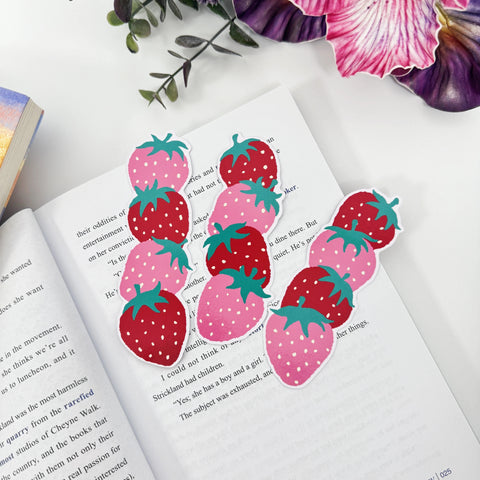Three red and pink strawberry bookmarks laid across an open book beneath pink and purple flowers