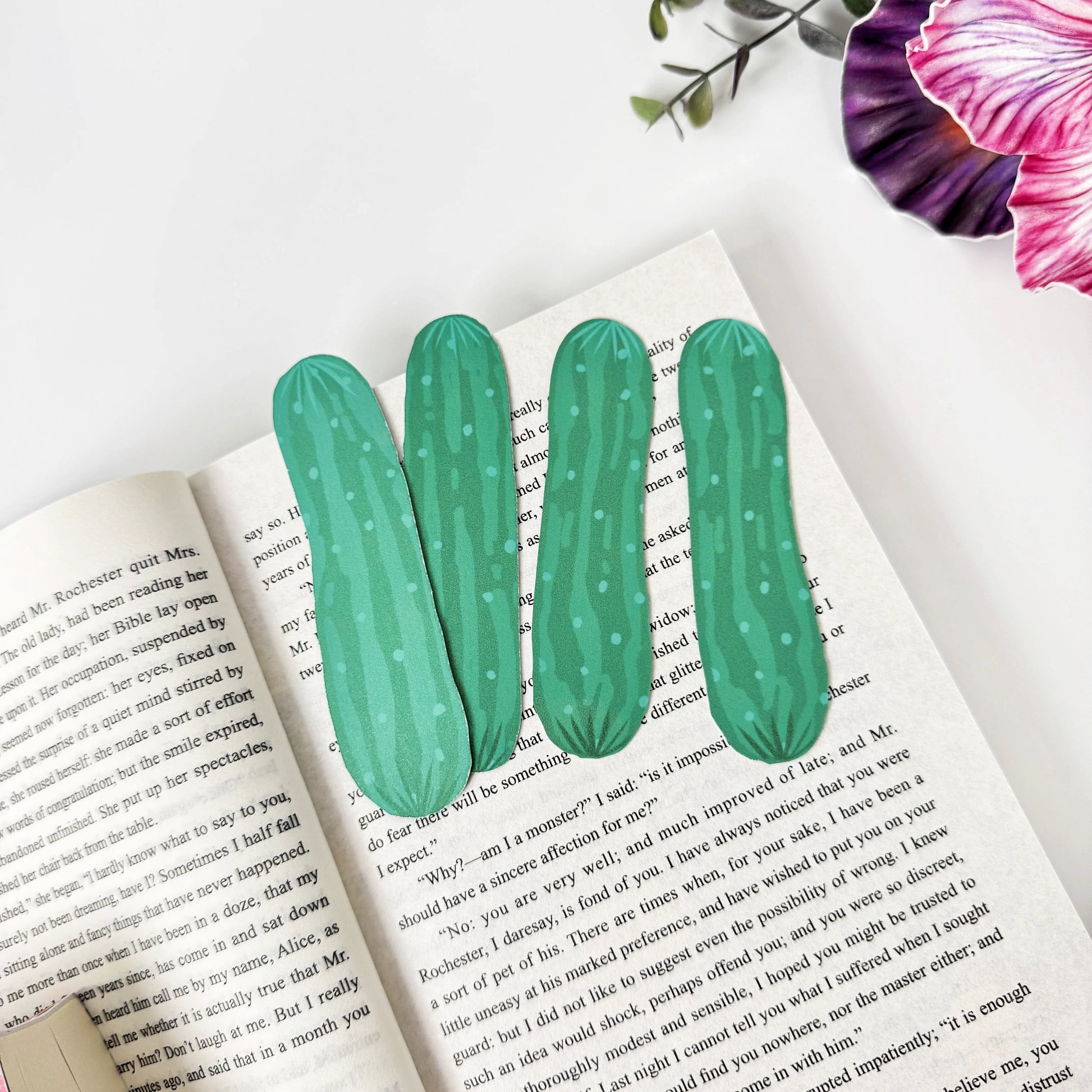 Four green cucumber bookmarks arranged across an open book beneath pink and purple flowers