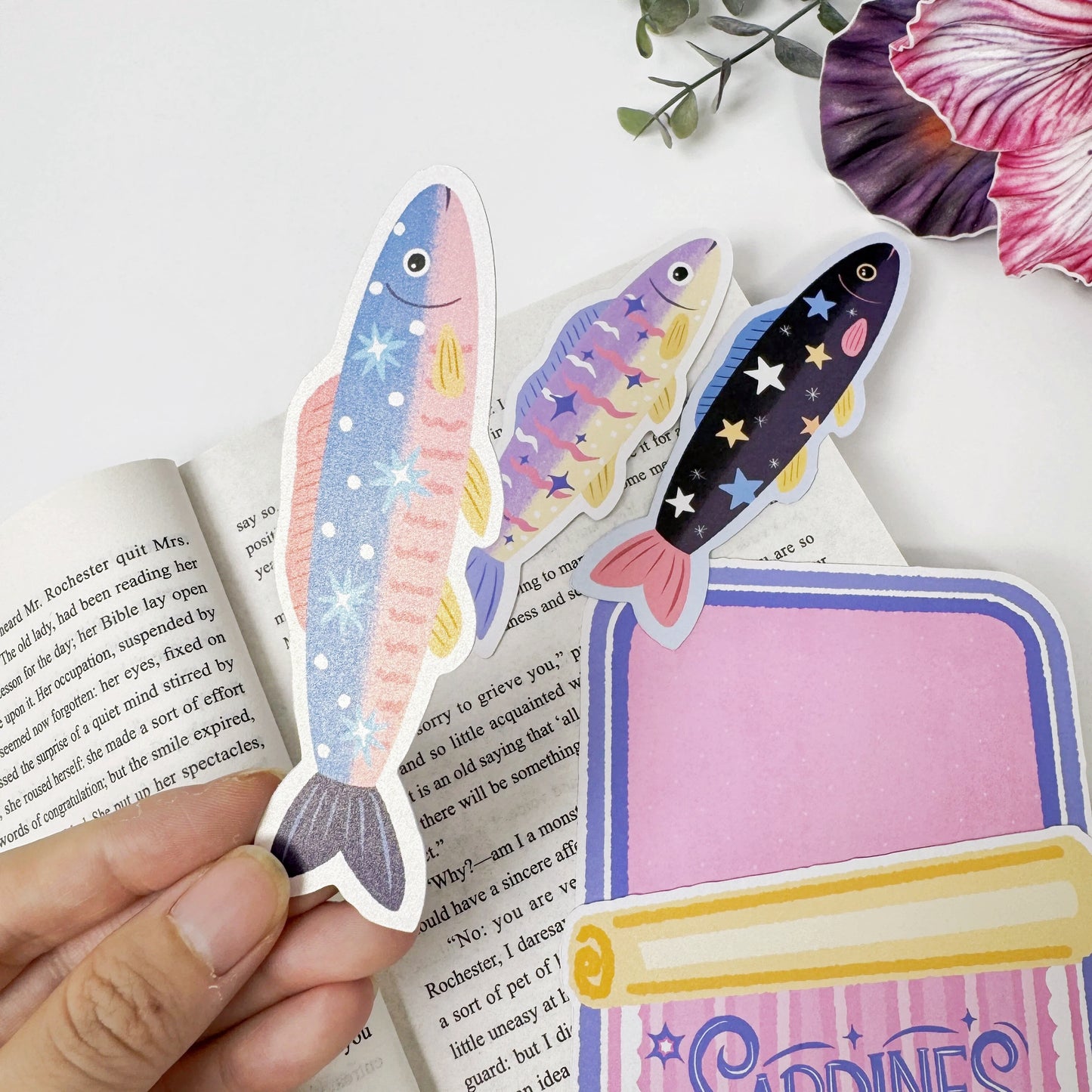 Close-up of a blue sardine bookmark held beside two fish bookmarks and the empty pink tin-shaped holder