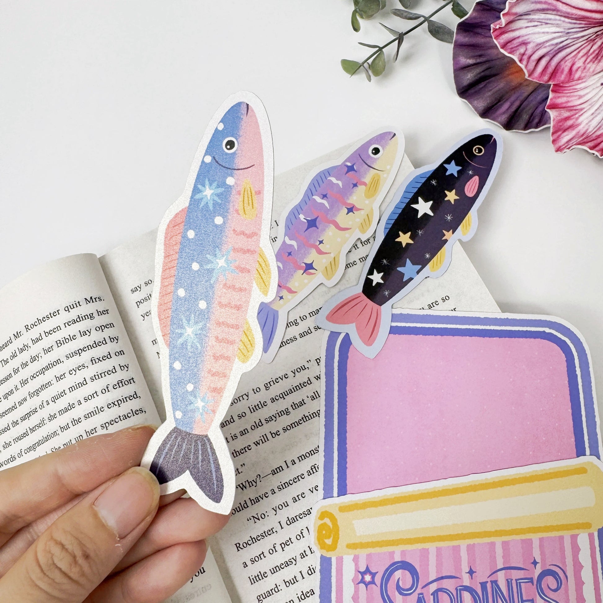 Close-up of a blue sardine bookmark held beside two fish bookmarks and the empty pink tin-shaped holder