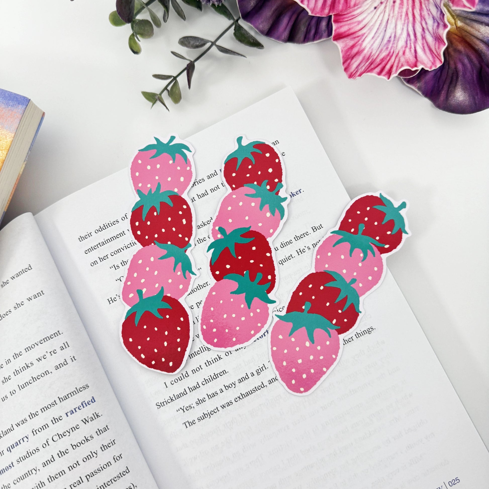 Three red and pink strawberry bookmarks laid across an open book beneath pink and purple flowers